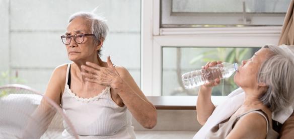 Twee vrouwen zitten in een huis voor de ventilator. Het is duidelijk warm. Een vrouw wappert met haar hand om af te koelen en de andere vrouw drinkt een glas water.